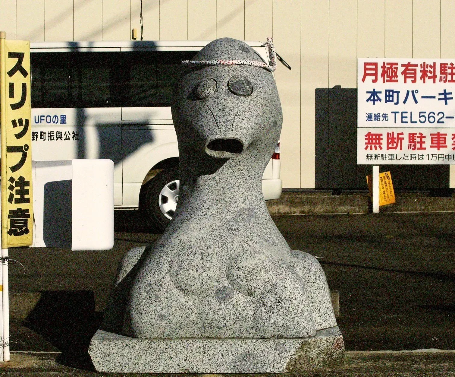 A stone sculpture of a creature with a long neck, small arms, and a face with closed eyes and an open mouth, wearing a hat with a string and small objects hanging from it, situated outdoors near a parking lot and Japanese signs.