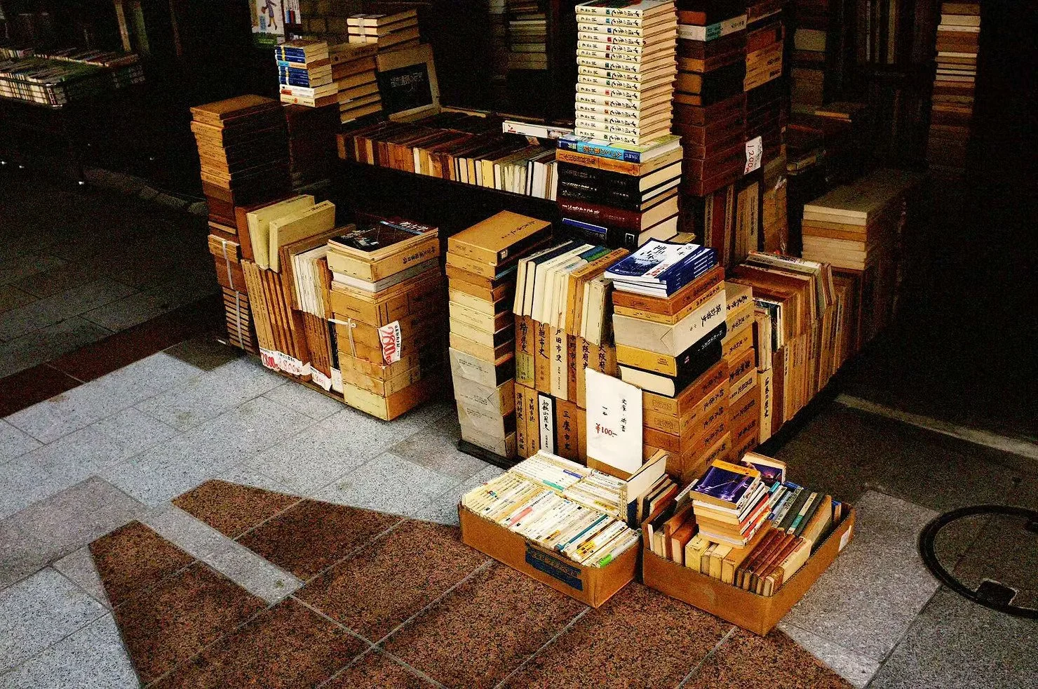 Stacks of books and magazines arranged on the ground and on a platform, with some books in cardboard boxes, near a tiled floor.