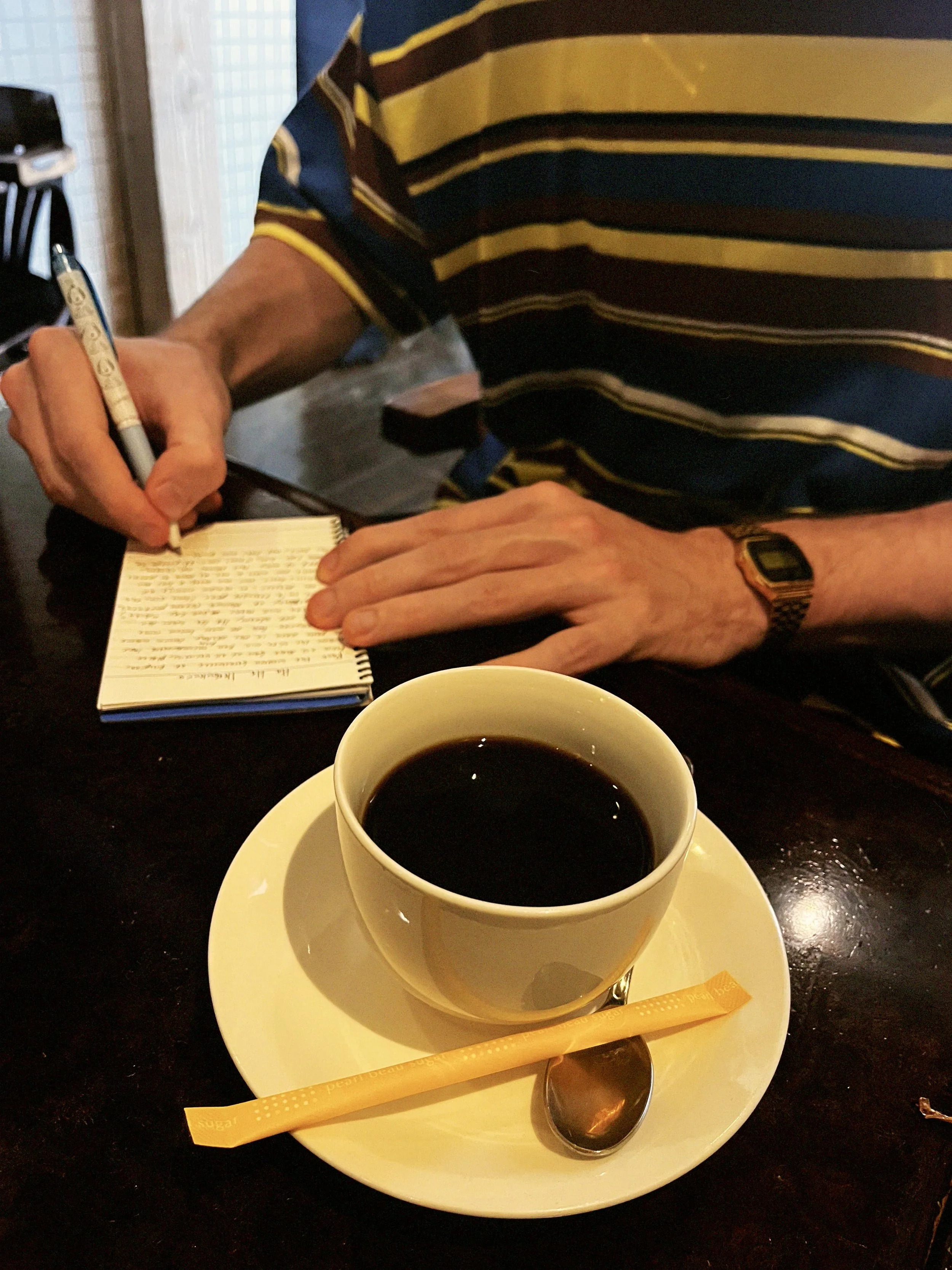 A person in a striped shirt sitting at a table, writing in a notepad with a pen. On the table in front is a white cup of black coffee on a saucer with a spoon and a sugar packet.