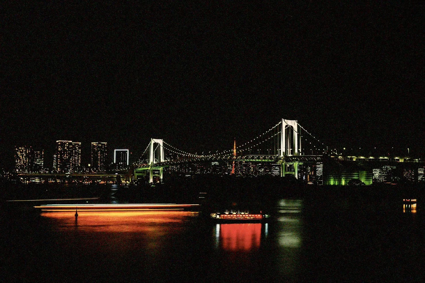 Night view of a illuminated city skyline with a large suspension bridge, water with reflections, and some light trails from moving boats or vehicles.