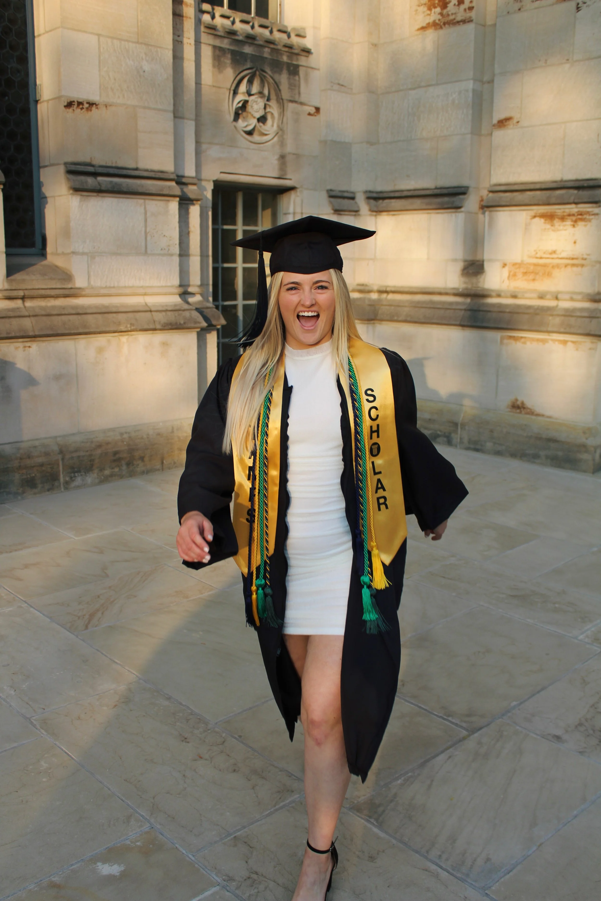 Young woman in a graduation cap and gown celebrating outdoors on stone pavement, with a historic stone building in the background.