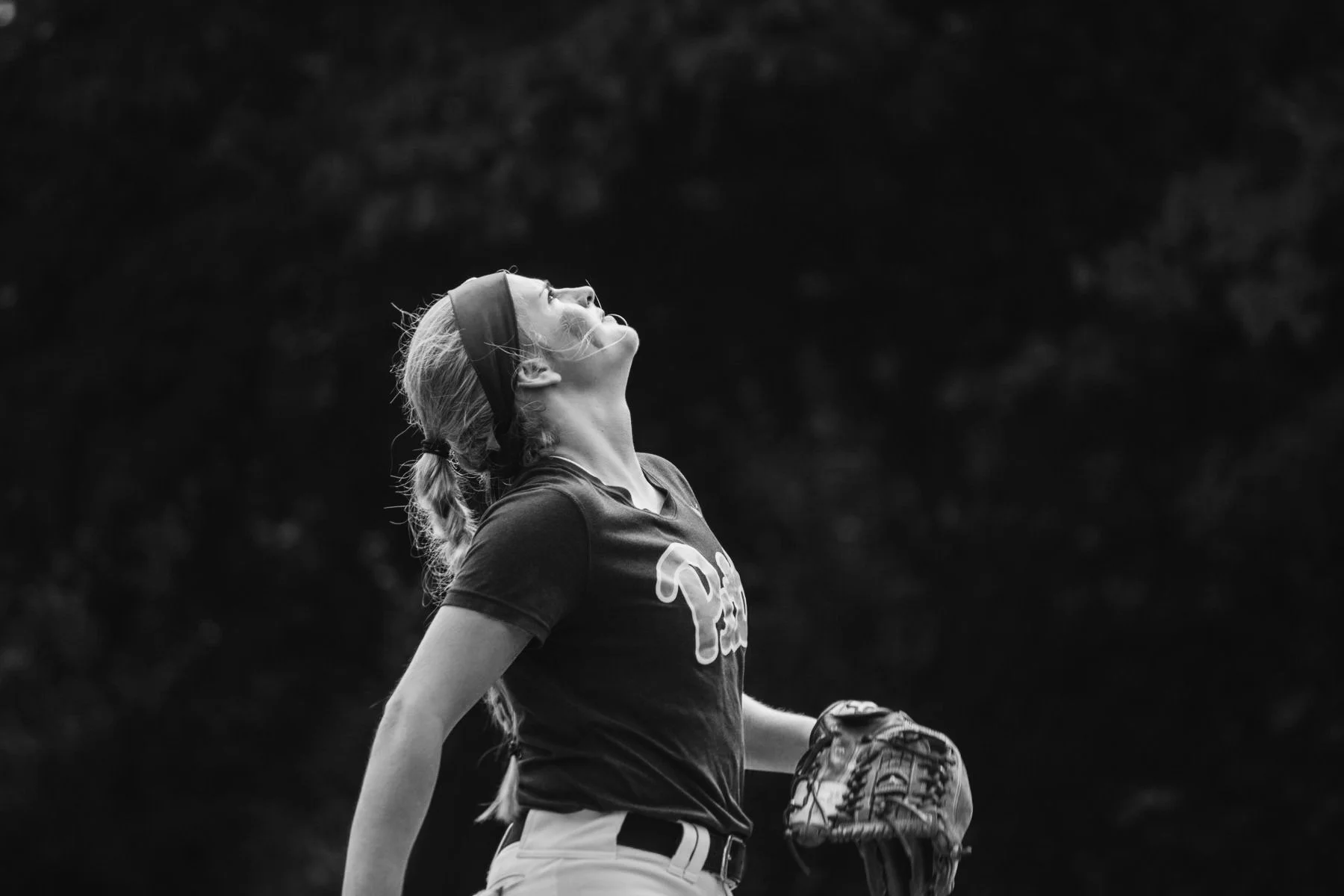 A young woman with blonde hair in a braid, wearing a headband and sports attire, stands outdoors holding a baseball glove, looking upwards.