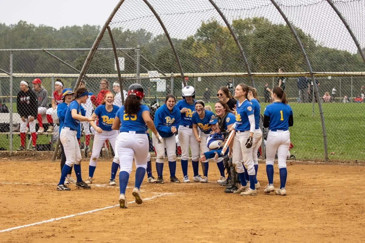 A women's softball team in blue uniforms celebrating on the field with their coach during a game, with spectators sitting behind a chain-link fence in the background.