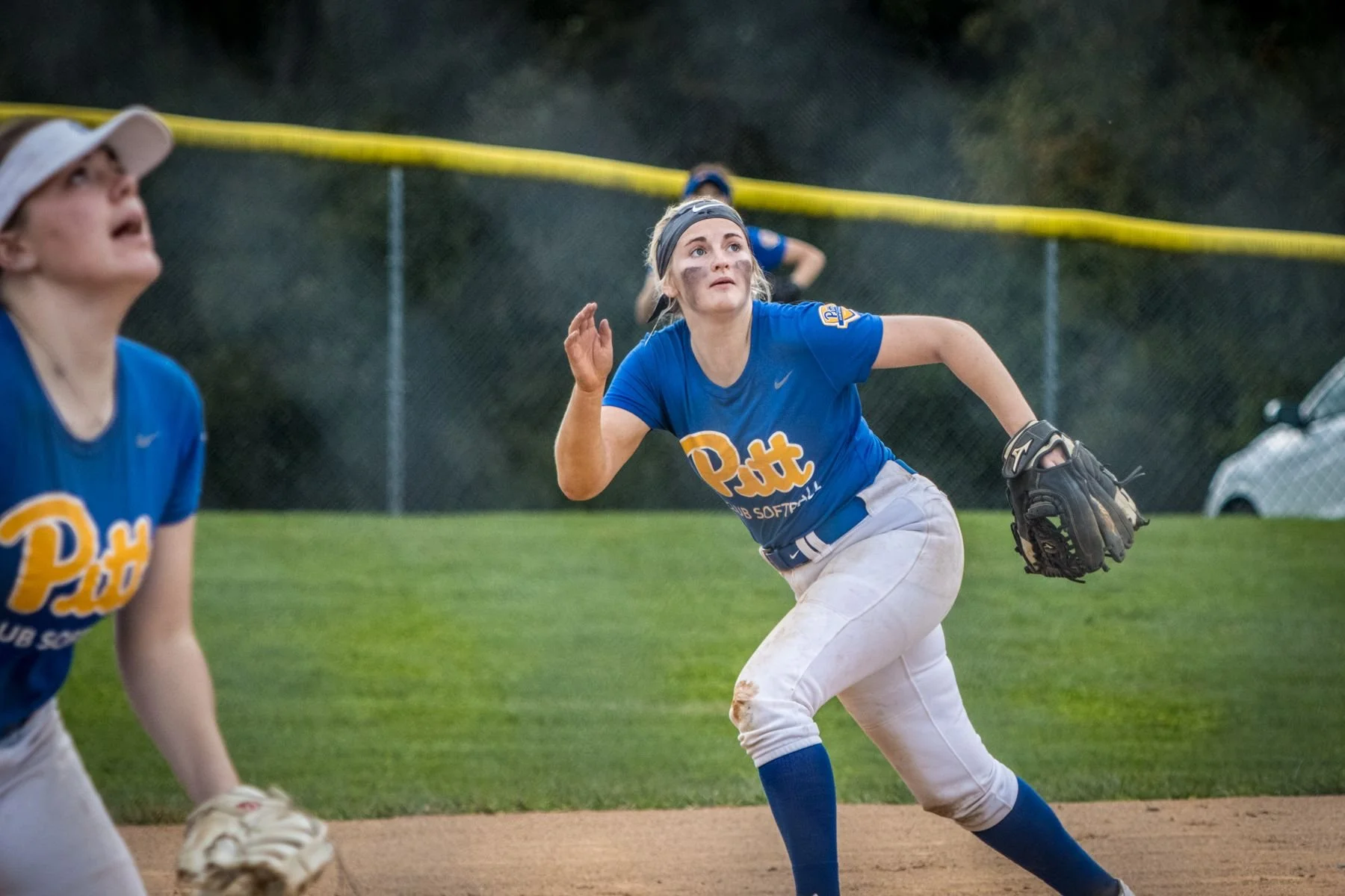 Softball players in blue uniforms playing on the field, with one player in a ready position and another preparing to catch a ball, during a game at twilight.