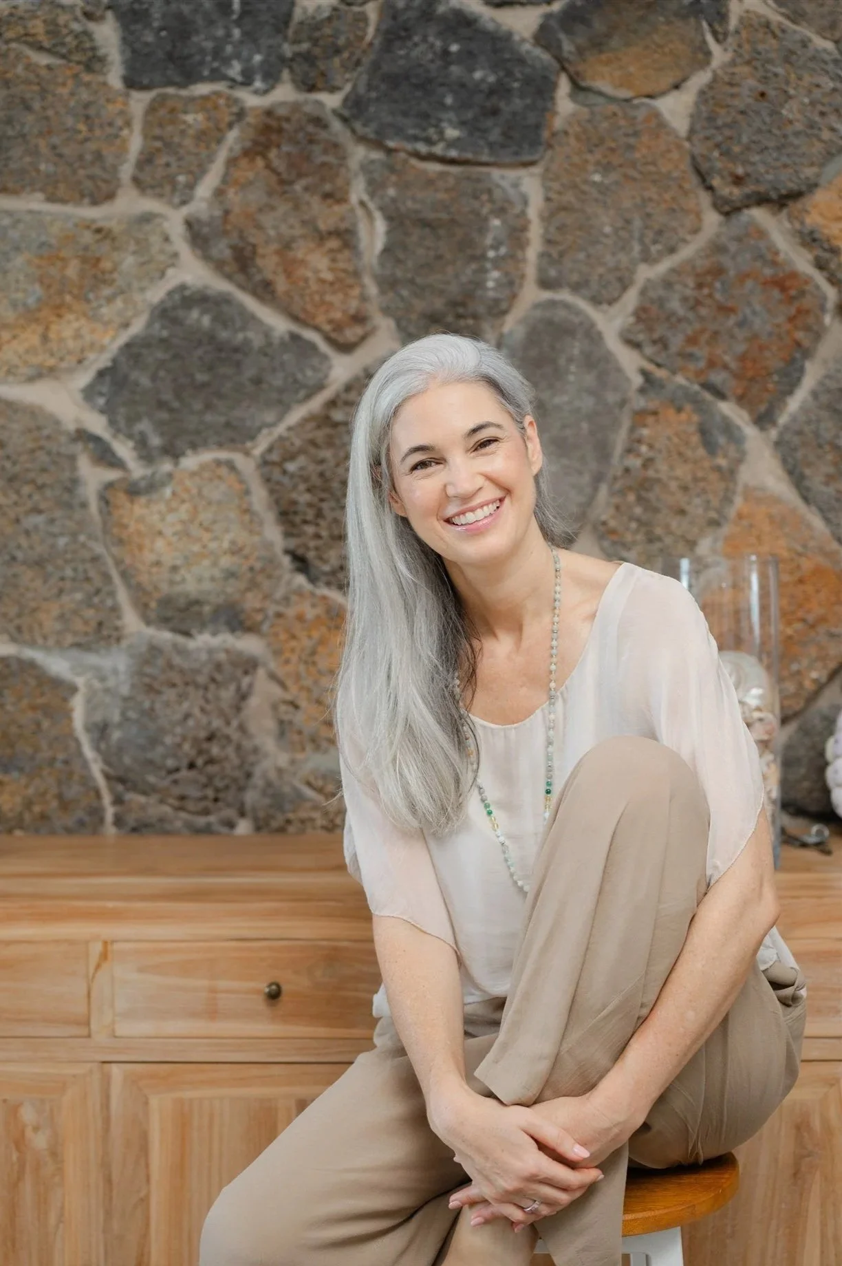 A smiling woman with long gray hair sitting on a wooden stool in front of a stone wall, wearing a light-colored top and beige pants.