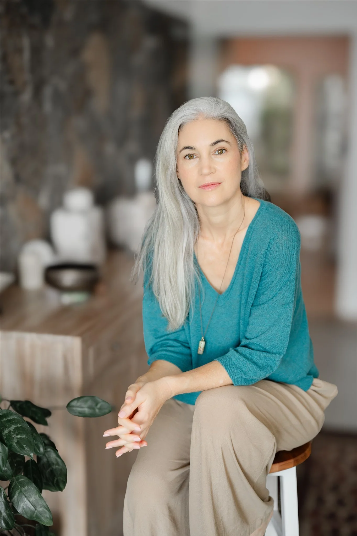 An older woman with long gray hair, wearing a teal long sleeve shirt and beige pants, sitting on a stool in a cozy indoor setting with a stone wall and blurred background, looking softly at the camera.
