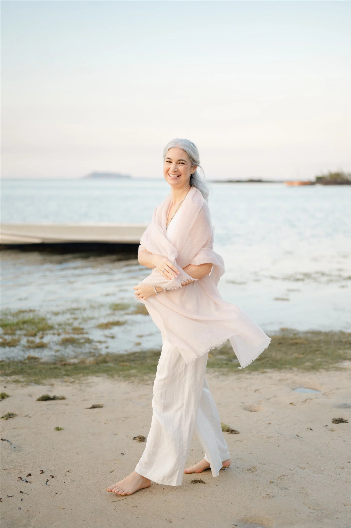 Stephanie Standinga t beach with the sea behind her with pink shawl looking at camera. Living with more ease, joy and abundance and flow.
