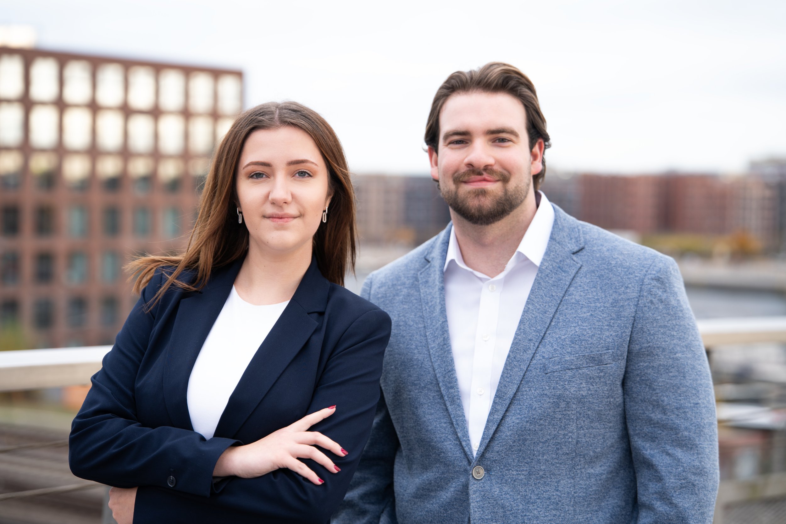 Ein Mann und eine Frau stehen auf einem Balkon im Freien mit einer städtischen Skyline im Hintergrund. Sie tragen formelle Kleidung, der Mann in einem blauen Blazer und die Frau in einem schwarzen Blazer.