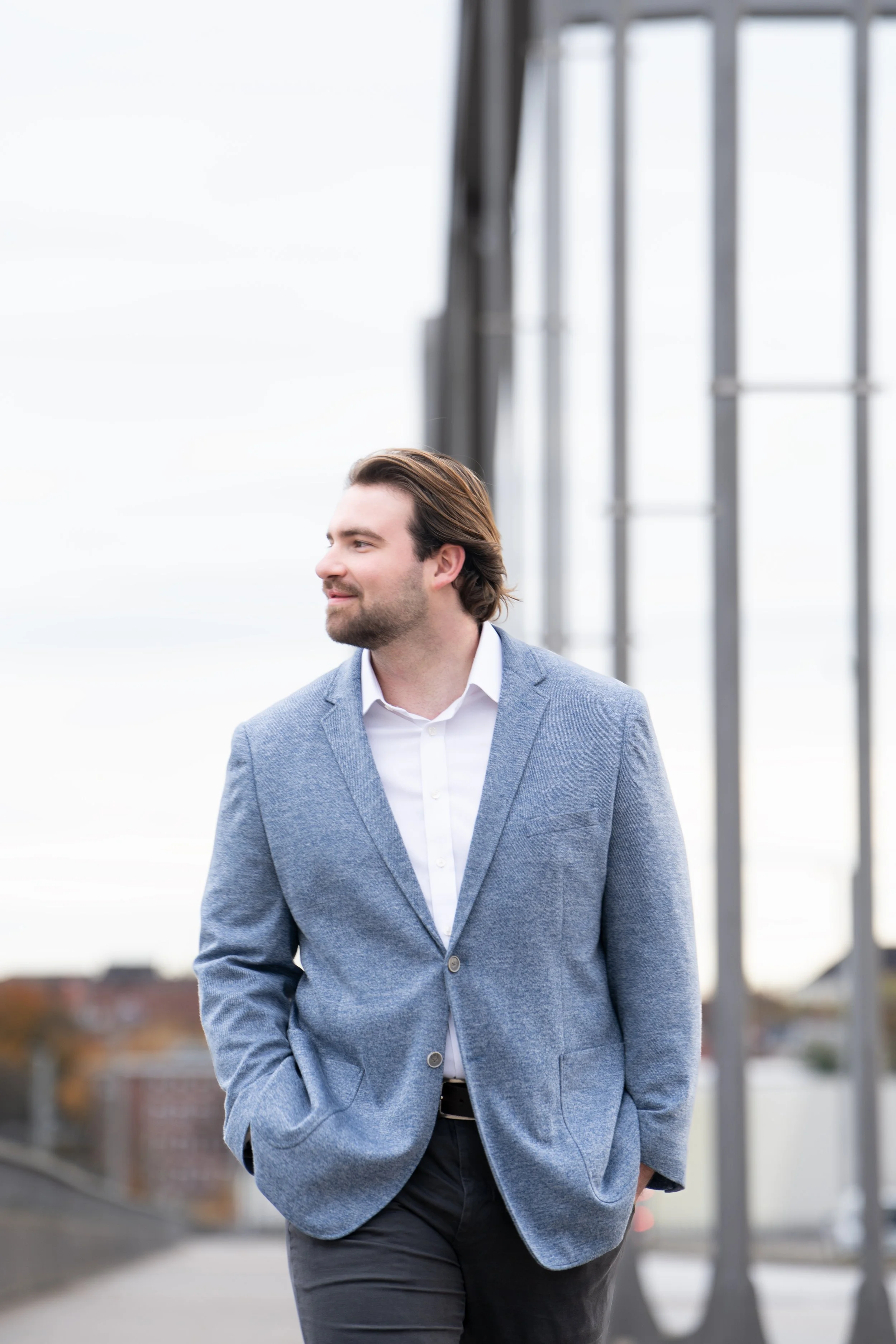 Ein junger Mann in einem grauen Blazer und weißen Hemd, der draußen auf einer Brücke steht und in die Ferne blickt.