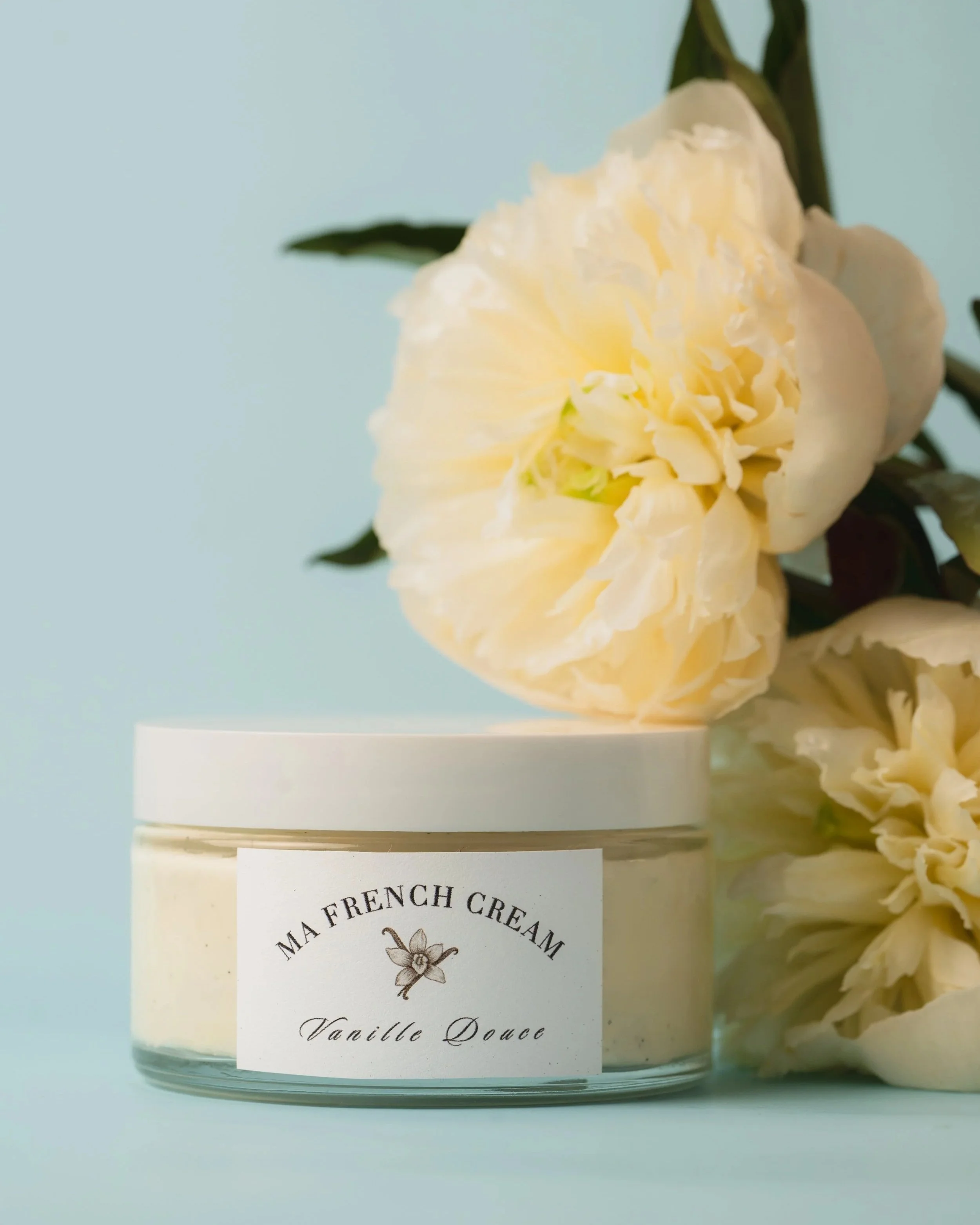 A jar of Ma French Cream Vanilla Douce with white lid, placed in front of light yellow peony flowers and dark green leaves against a pale blue background.