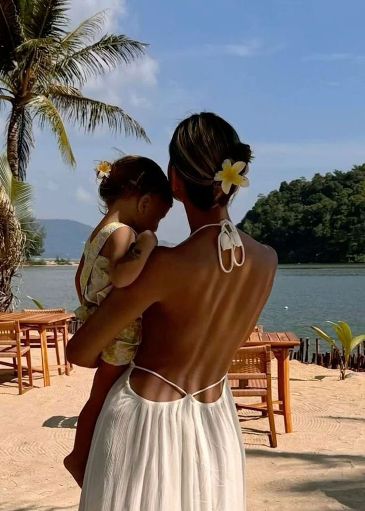 A woman holding a small child on a beach with palm trees, wooden chairs, water, and a small island in the background. Both have flowers in their hair.