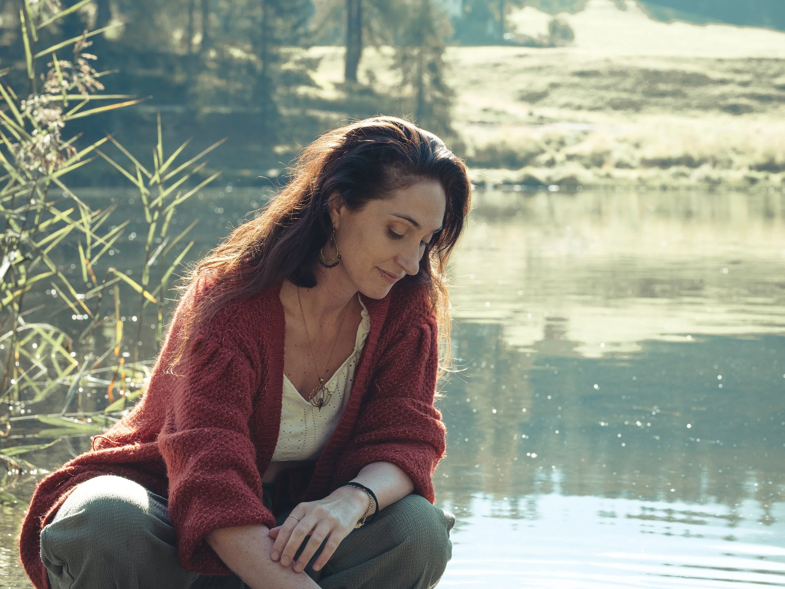 Ein junge Frau mit langen braunen Haaren sitzt am Fluss, trägt eine rote Strickjacke und schaut nach unten, während die Natur im Hintergrund sonnig ist.