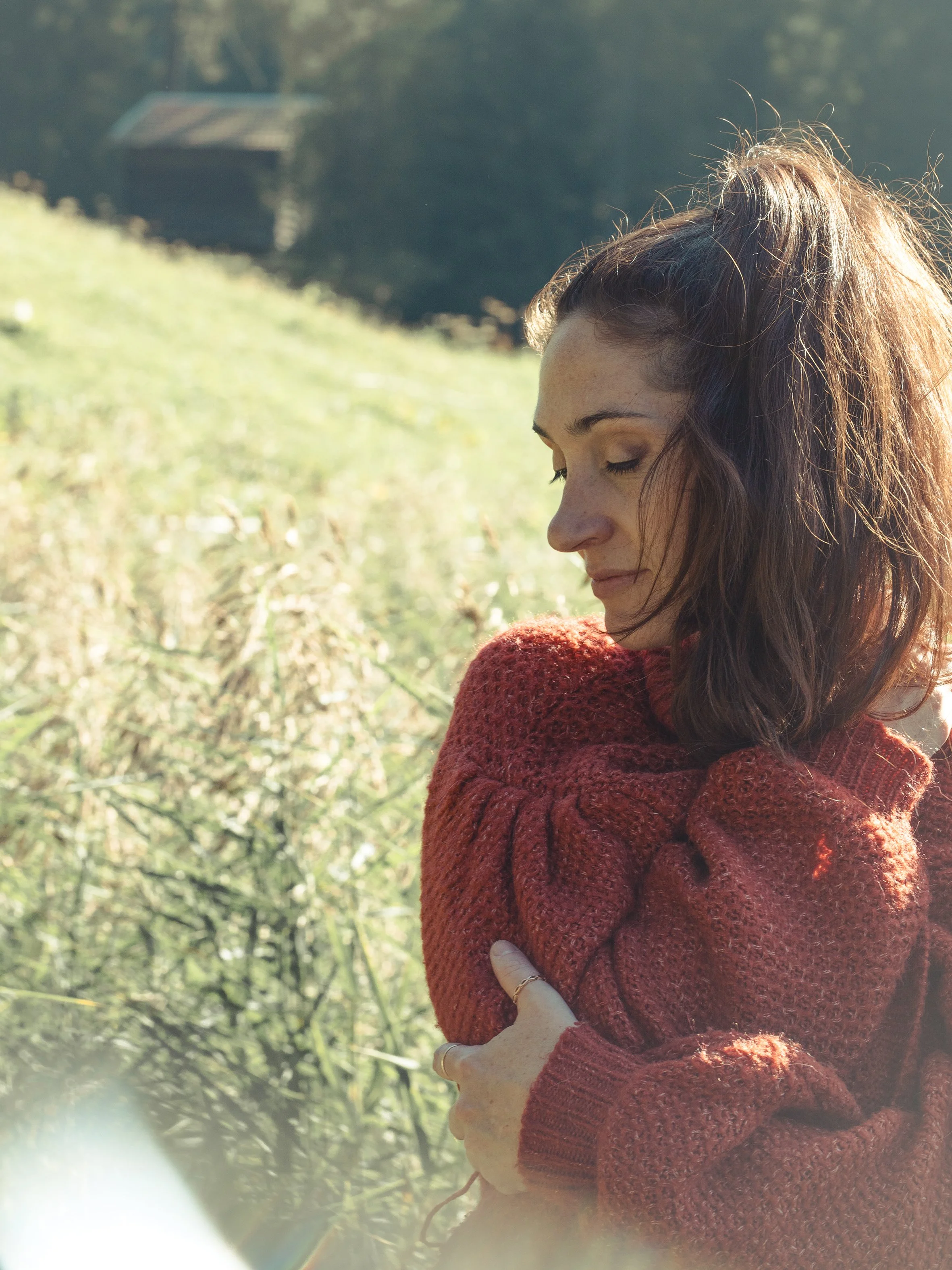 Eine junge Frau trägt einen roten, gestrickten Pullover, steht im Freien und hat die Arme verschränkt. Sie hat braune, gewellte Haare und ist im Profil zu sehen, mit geschlossenen Augen.