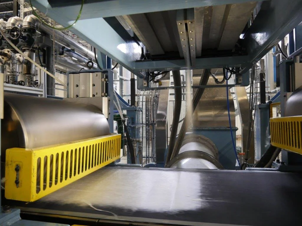 Industrial manufacturing equipment with metal rollers, pipes, and a conveyor system in a factory setting.
