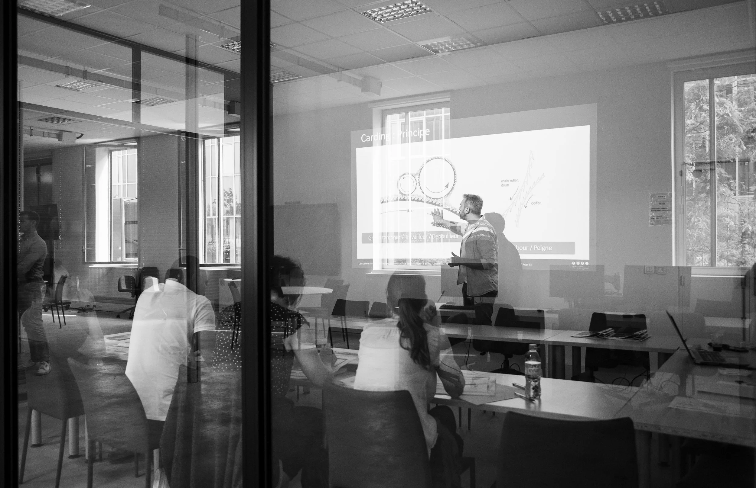 A classroom or conference room with a man giving a presentation to a group of students or attendees, visible through a glass window. The presenter is standing in front of a projected slide with diagrams and text, gesturing with his hand. Several people are seated around tables, some taking notes, and a water bottle is on one of the tables. Large windows allow natural light into the room.