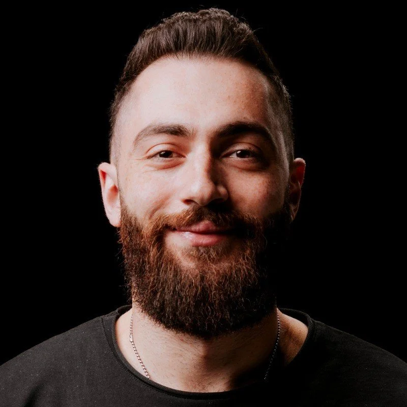 A young man with a beard and mustache, smiling, against a black background.
