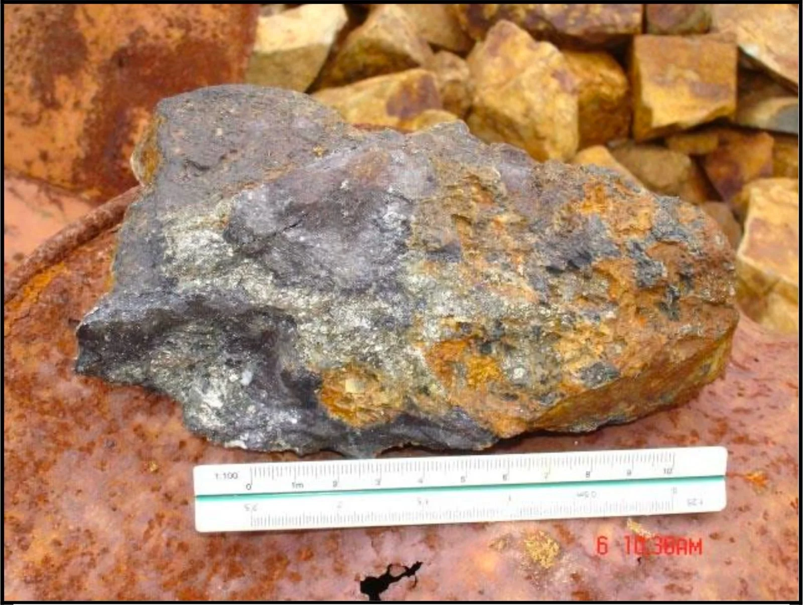 A large, multicolored rock with a rough surface, placed on a reddish-brown surface, with smaller rocks in the background. A measuring scale is positioned below the rock, showing centimeters and inches.