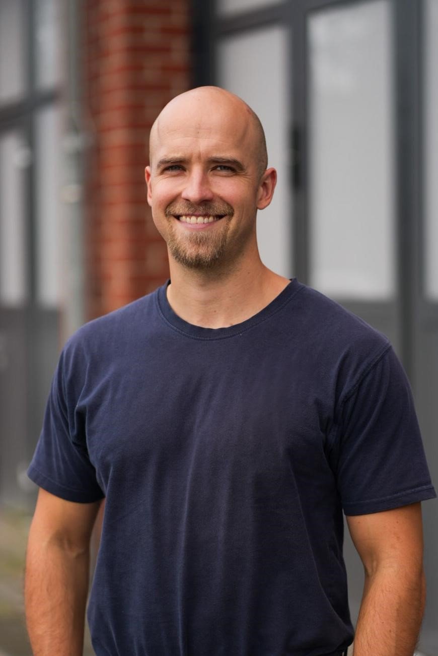 Ein lächelnder, glatzköpfiger Mann trägt ein dunkelblaues T-Shirt, steht vor einem modernen Gebäude aus Glas und Ziegel.