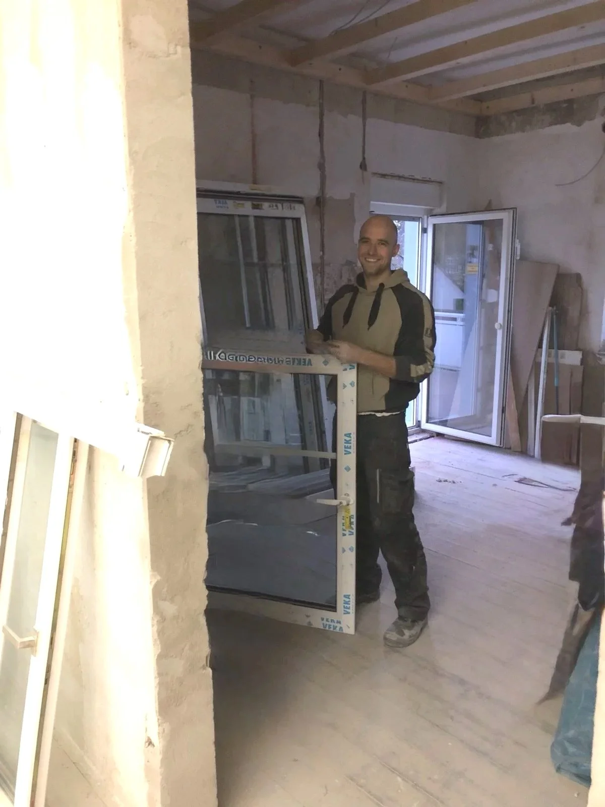 Ein Mann steht in einem Raum unter Bauarbeiten, hält ein Fenster in den Händen und lächelt. Im Hintergrund sind weitere Fenster und Baumaterialien sichtbar.