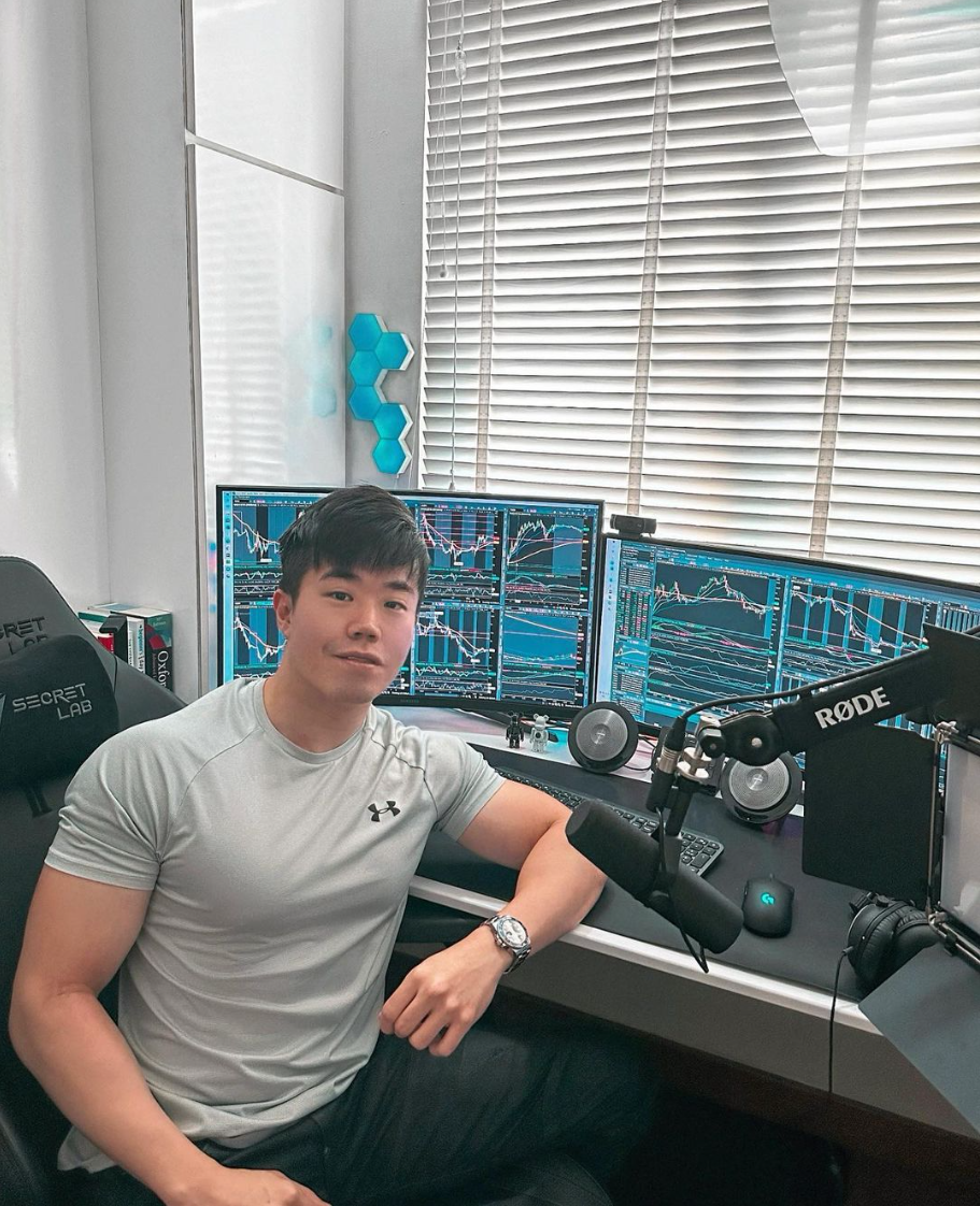 A young man in a light gray Under Armour shirt sitting at a desk with multiple monitors displaying stock charts, in a room with white blinds and blue hexagonal wall decorations. There is a professional microphone, speakers, and a webcam on the desk.