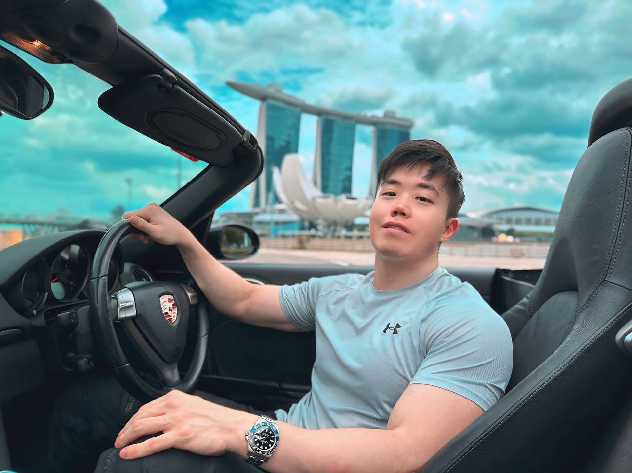 Young man in a gray Under Armour t-shirt sitting in a black Porsche convertible with the city skyline and Marina Bay Sands hotel in the background.