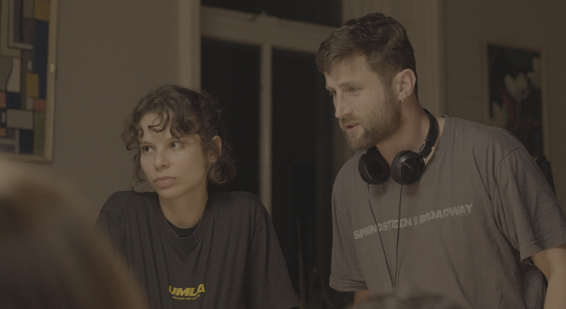 Two individuals engaged in a discussion. The person on the left, a woman, has short curly hair and is wearing a black shirt with yellow text. The person on the right, a man, has short hair, a beard, and is wearing a gray shirt with white text and black headphones around his neck.