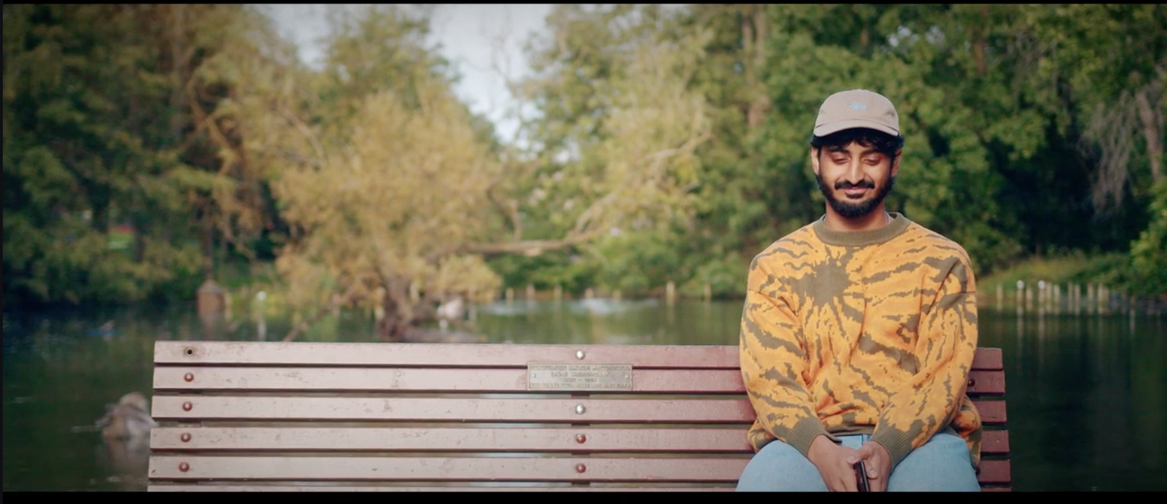 A man with a beard and mustache in a beige cap and yellow camo sweater, sitting on a park bench near a river with green trees in the background, smiling with closed eyes.