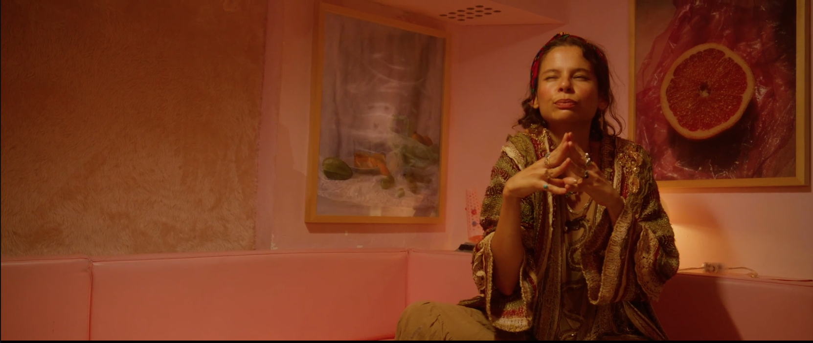 A woman with curly hair sitting on a pink sofa with her hands clasped together, resting her chin on her fingers, and looking into the distance. The room has pink and orange lighting, with framed art on the wall, including a photograph of a grapefruit and another image with objects and smoke.