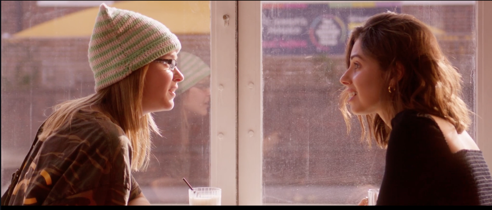 Two women sit across from each other inside a cafe, engaging in conversation. One wears a colorful knit beanie and glasses, the other has shoulder-length wavy hair and a black top. A glass of milk with a straw is on the table.