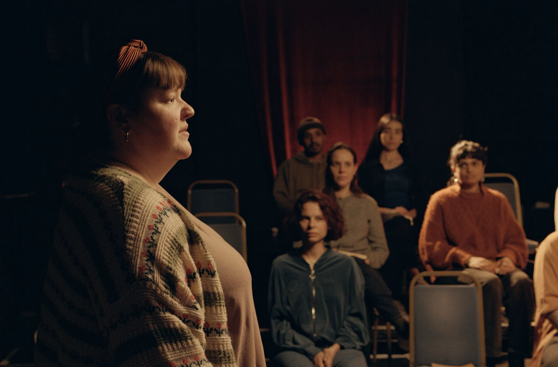 A woman with red hair and a patterned cardigan stands in profile in the foreground, while five other people sit or stand behind her in a dimly lit room with red curtains.