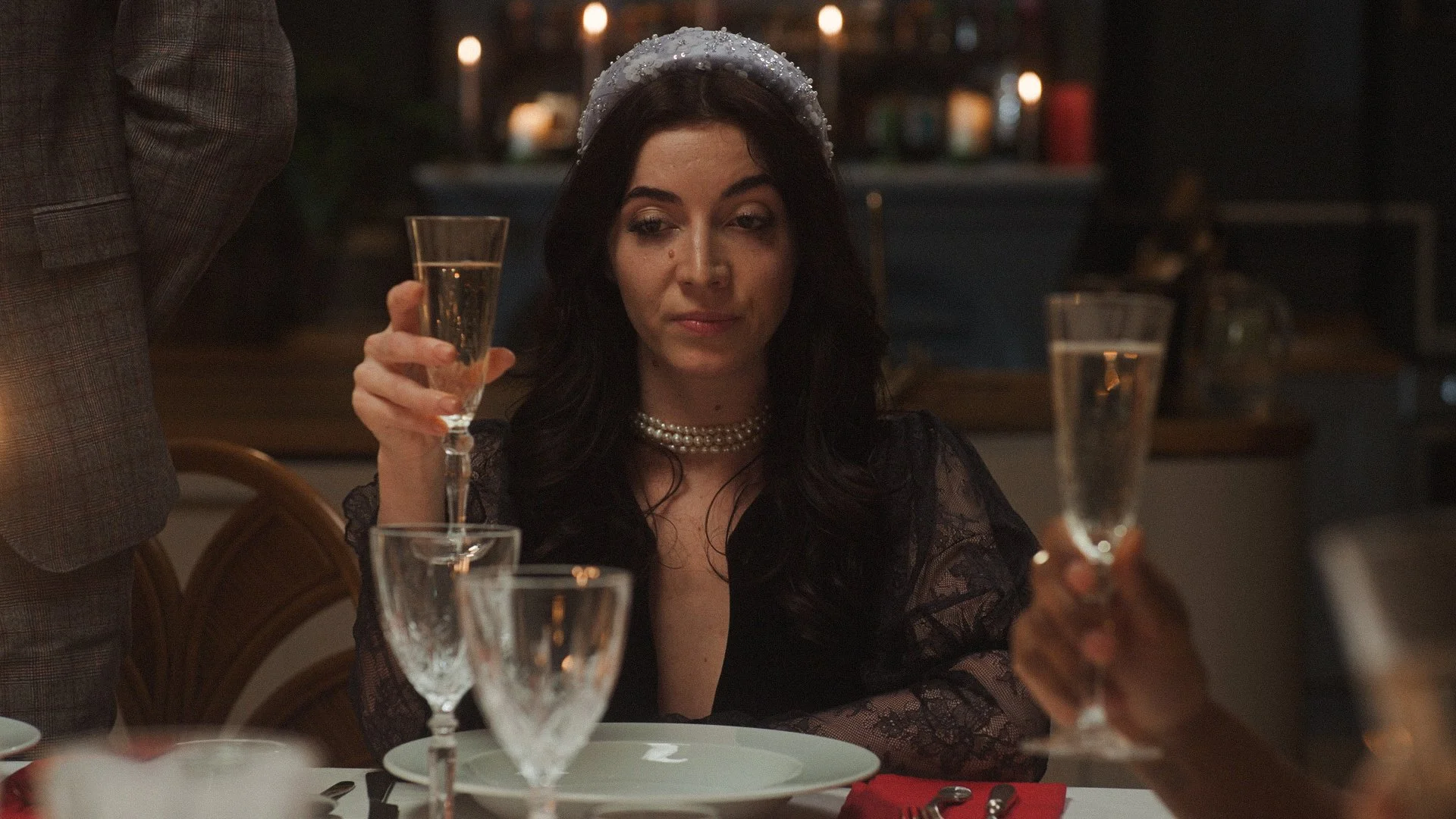 A woman with dark hair wearing a pearl necklace and a headband at a dinner table, holding a glass of champagne.