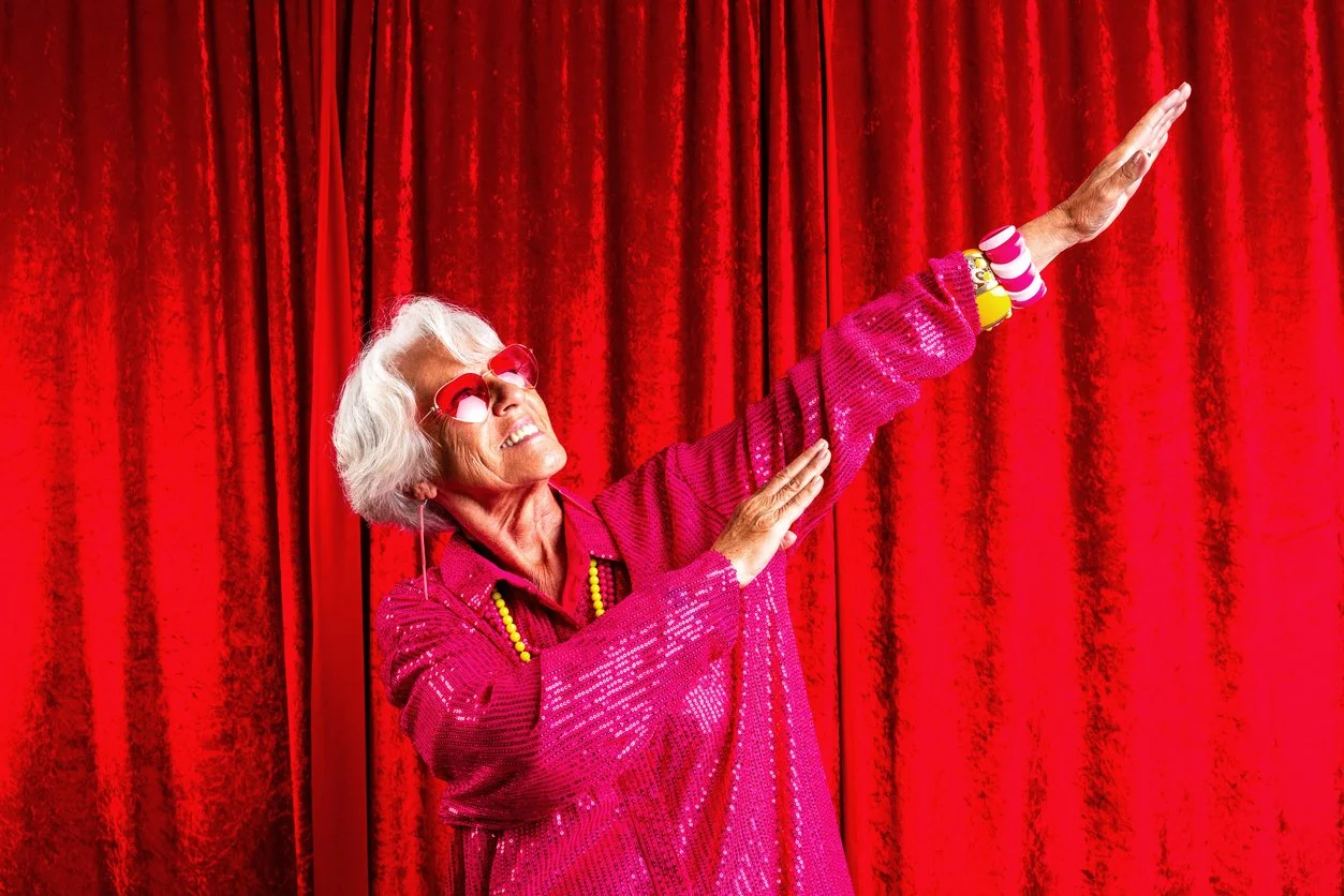 En eldre kvinne i rosa med solbriller og fargerike armbånd, danser foran en rød teppebakgrunn.