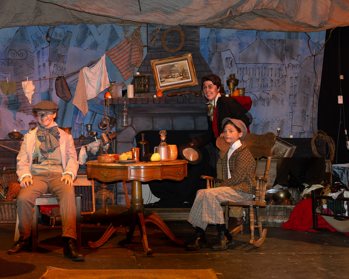 Skuespill på scenen med tre personer i tidstypiske klær, foran en peis, dekorert med gamle gjenstander og malerier, i en setting som etterligner en gammel bolig.