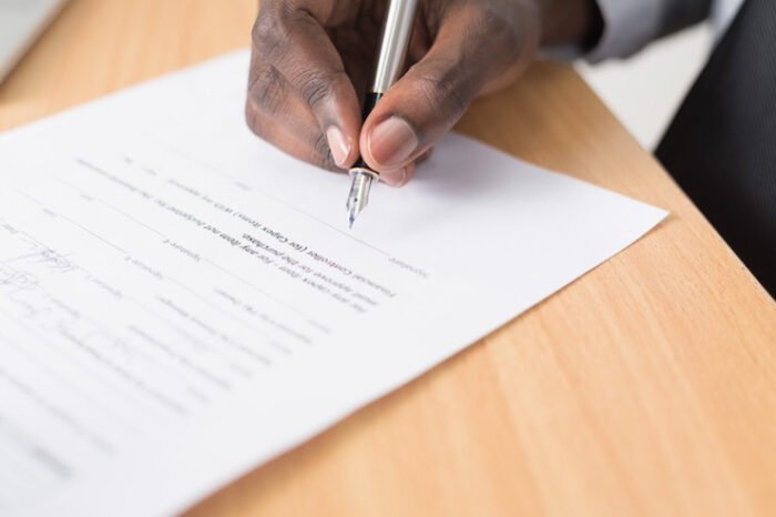 A person with dark skin is signing a document with a silver pen on a wooden desk.