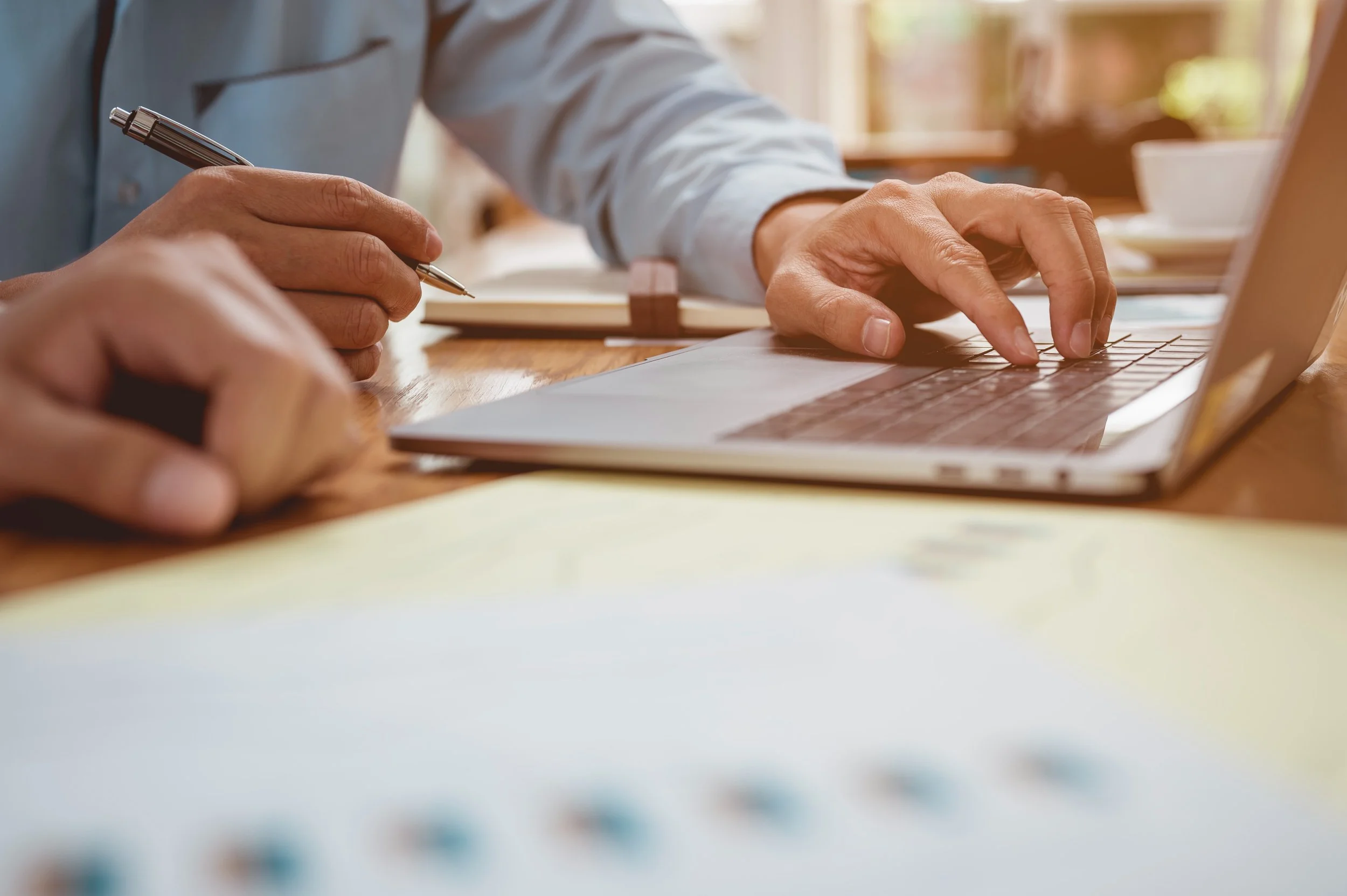 Hands of two people working at a wooden table, one typing on a laptop and the other holding a pen near a notebook.