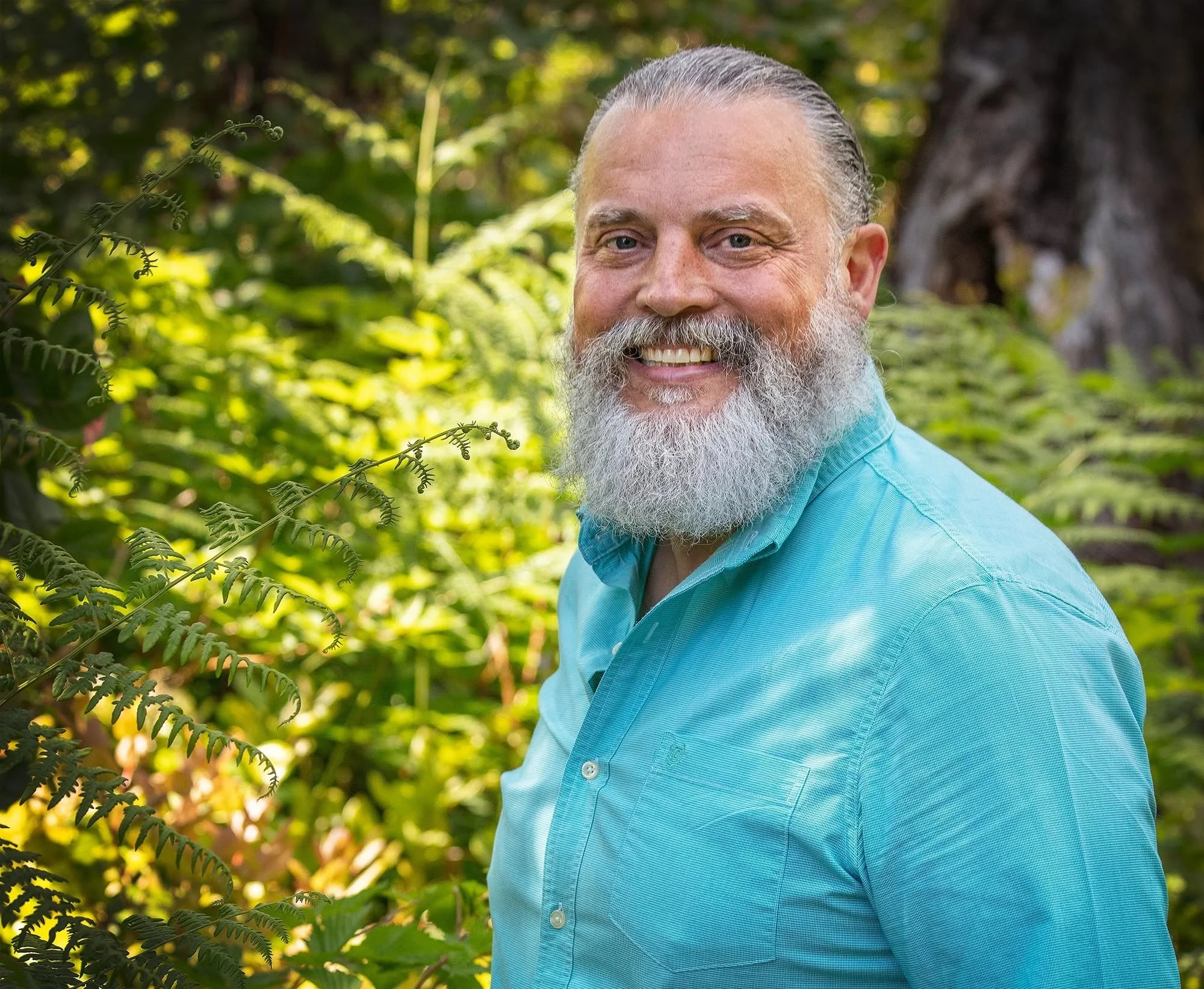 Portrait of a smiling middle-aged man with a white beard and long hair slicked back, wearing a light blue button-up shirt, standing outdoors with green foliage and a tree in the background.