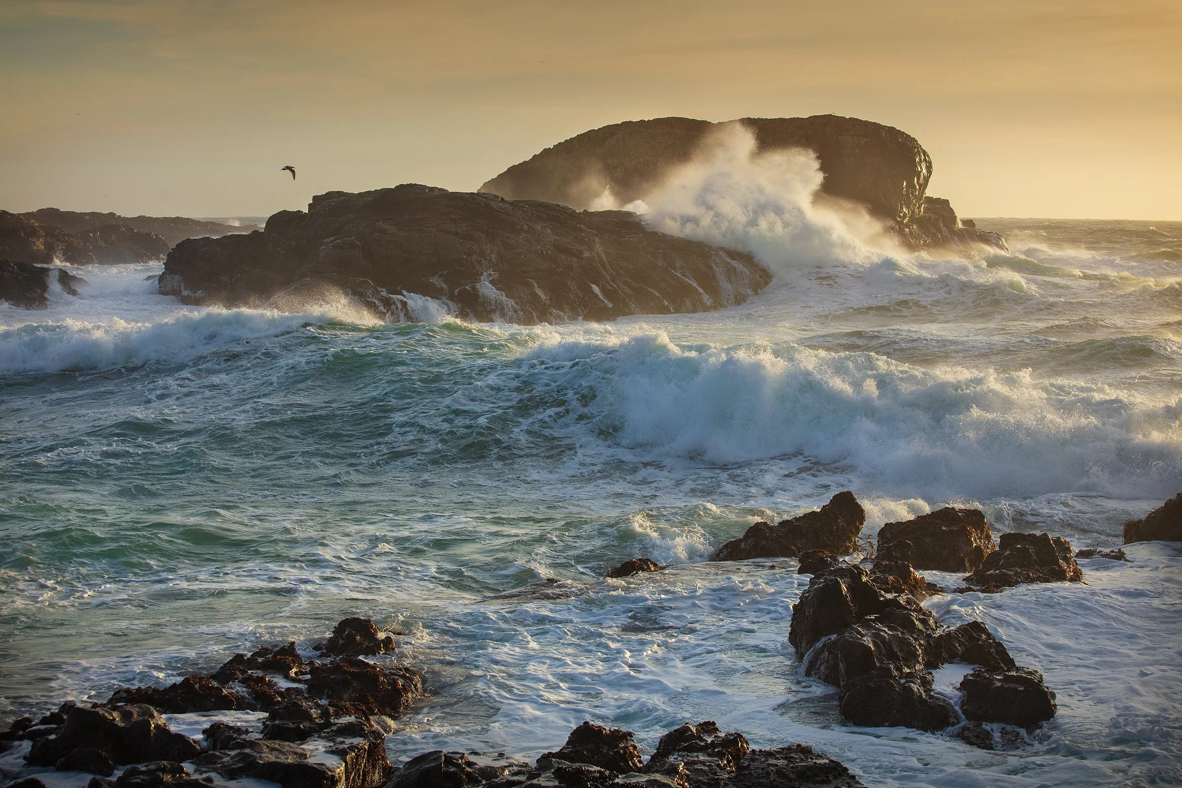 Ocean waves crashing against rocks with a large island or rock formation in the background, during sunset or sunrise.