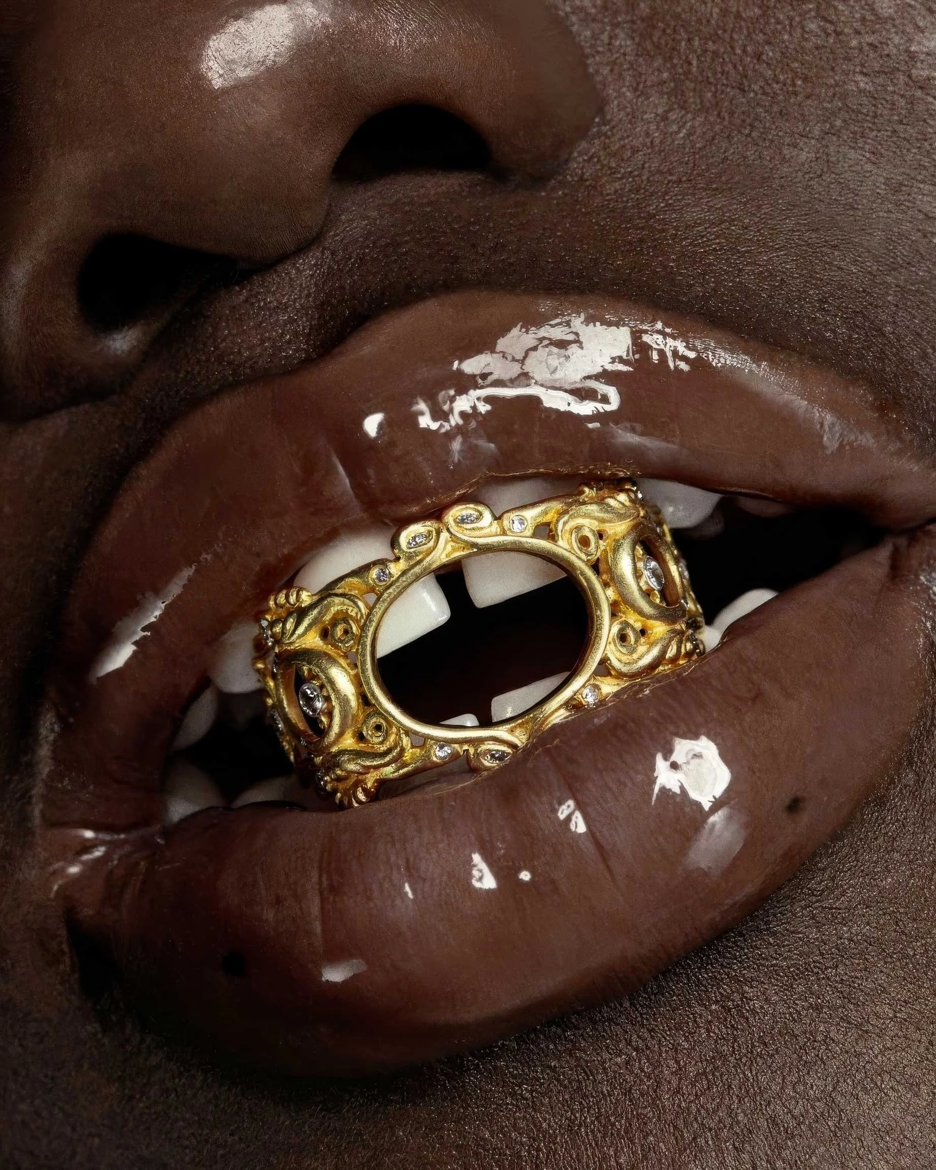 Prima piano di un volto con labbra carnose, che portano un anello d'oro con dettagli elaborati e pietre preziose, inserito tra i denti.