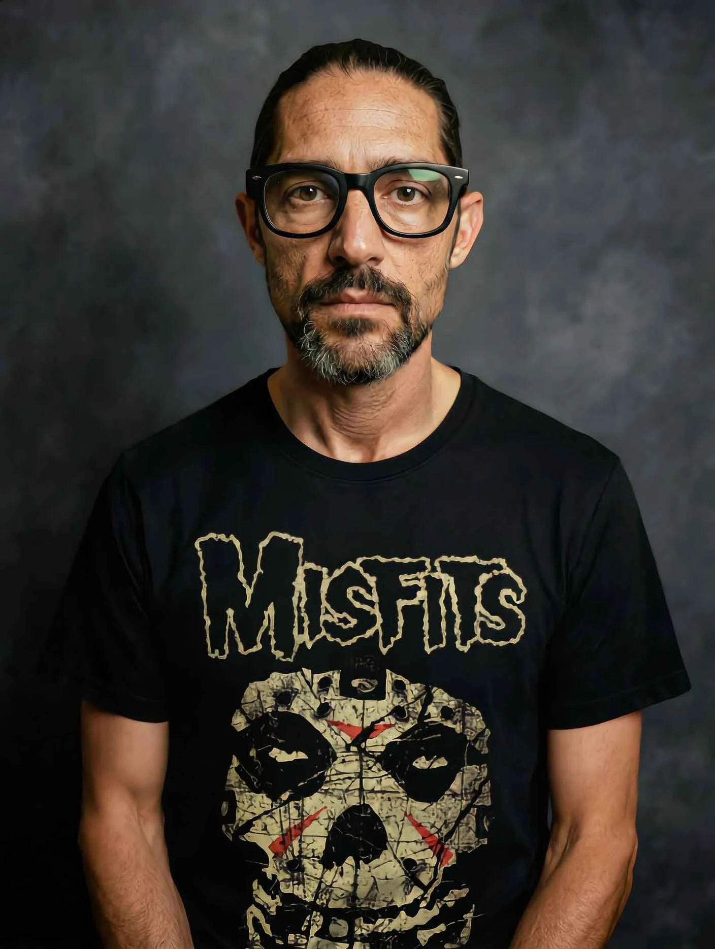 ritratto di un uomo con occhiali spessi, barba e capelli lungi, vestito con maglietta dei Misfits con un teschio. sfondo scuro