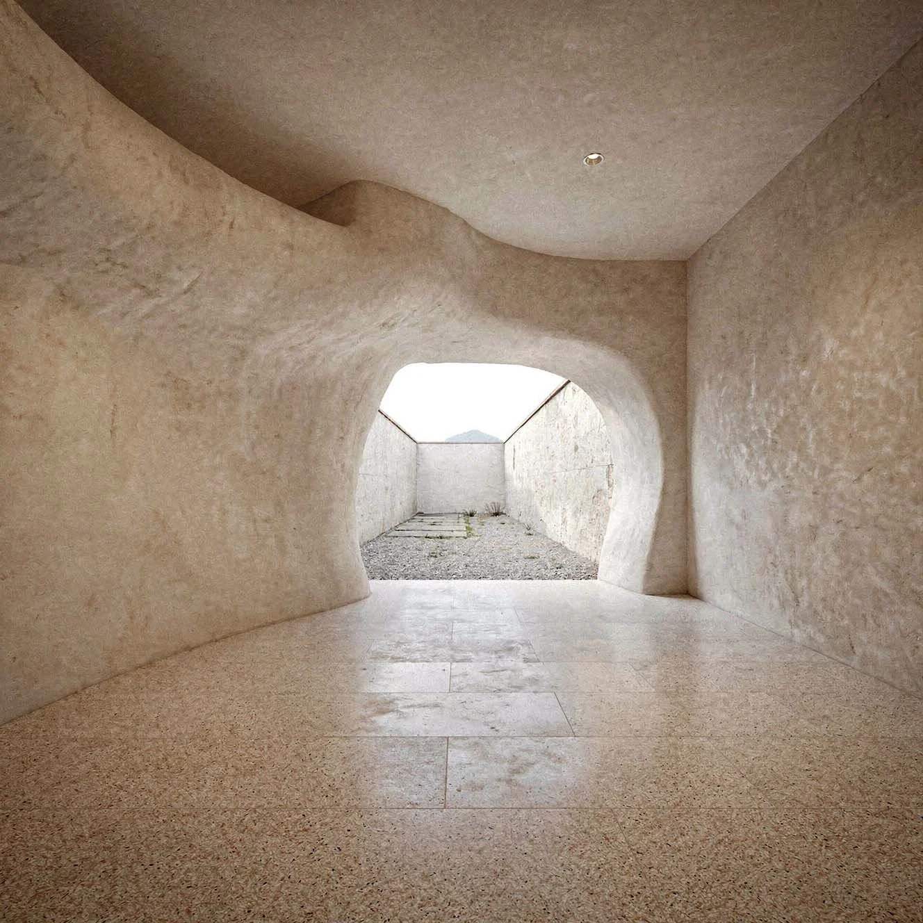 Interno di una struttura con pareti curve in pietra e un'apertura che mostra un cortile esterno con muro bianco e montagne in lontananza.