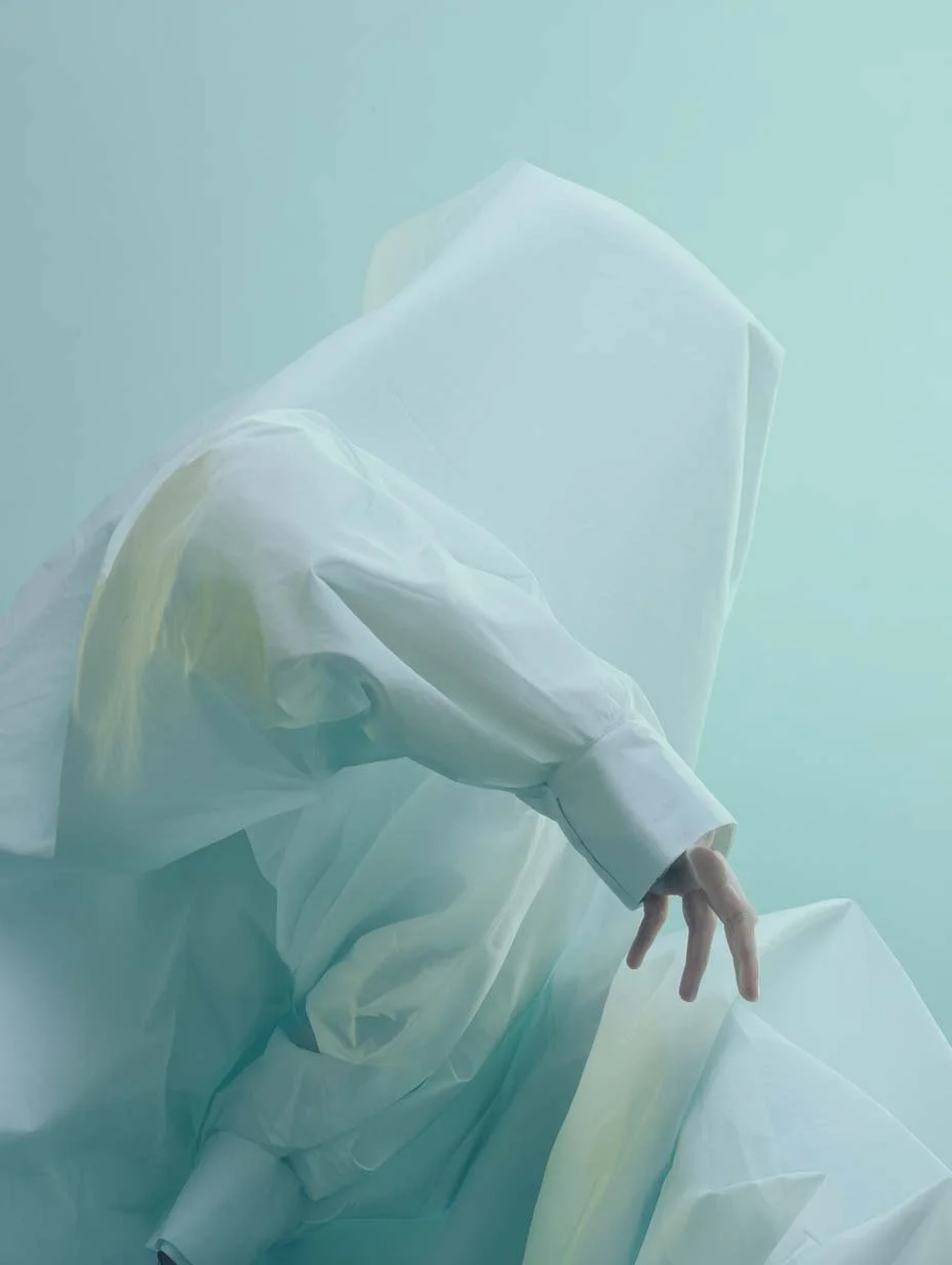 Persona vestita con abbigliamento bianco con volto nascosto, seduta su un oggetto bianco, con una mano che si piega in modo artistico.