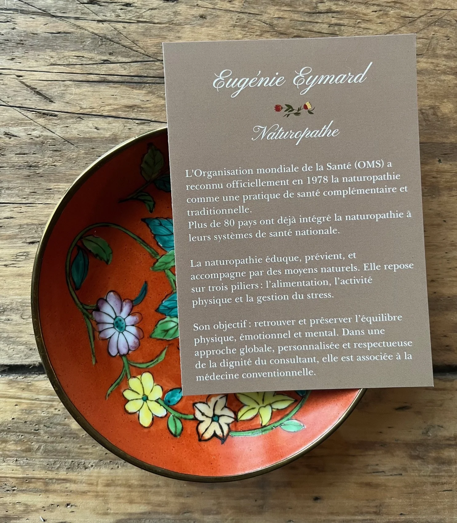 A decorative bowl with colorful floral patterns inside, placed on a wooden surface. A gray informational card about naturopathy is resting on the bowl.