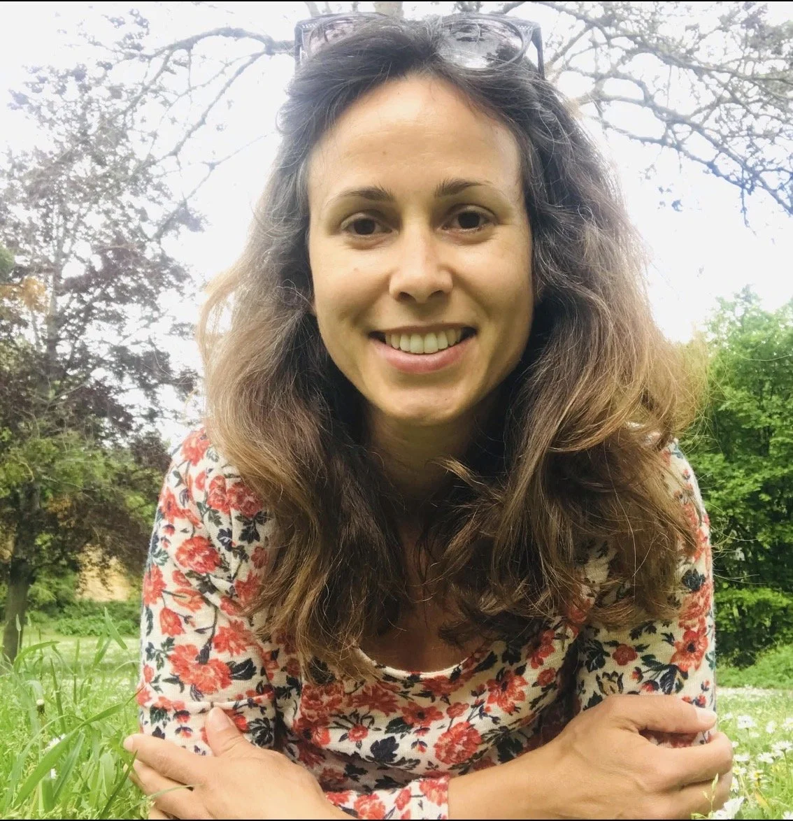 A woman with long, wavy brown hair and glasses resting on her head, smiling outdoors among trees and greenery.