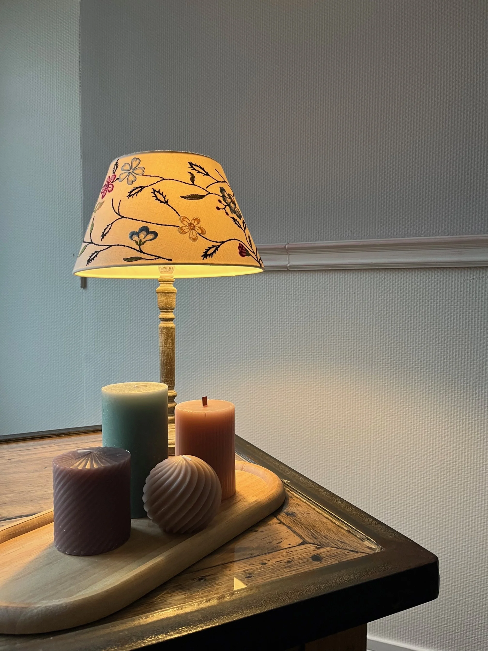 Decorative table lamp with floral patterned lampshade, set of four candles on a wooden tray, and a wooden table.