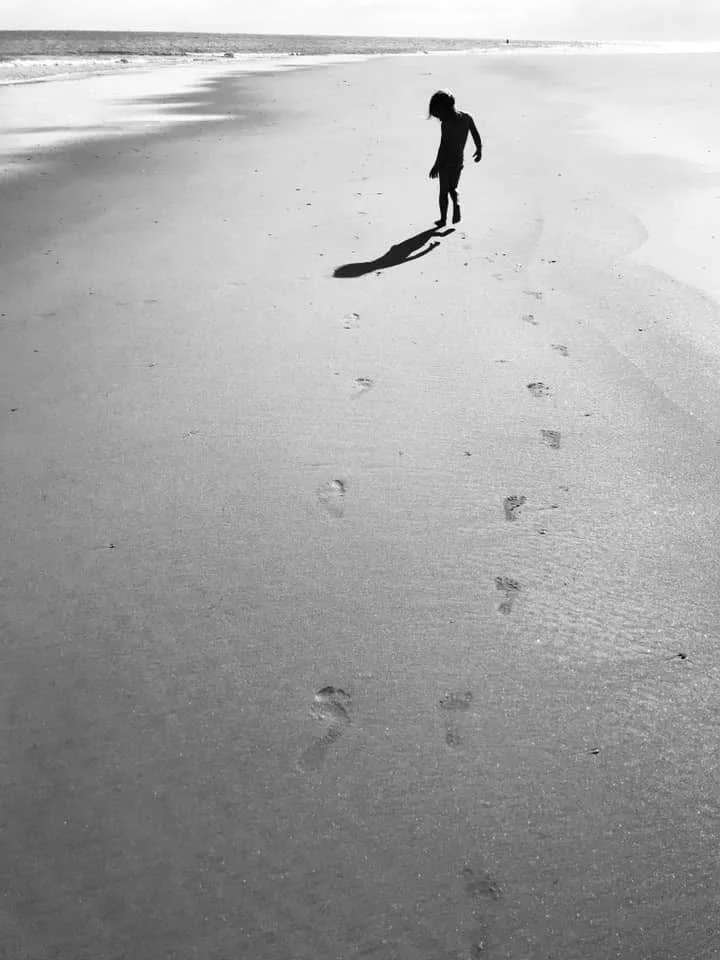 A child walking alone on a sandy beach, casting a long shadow, with footprints behind them and the ocean in the background.