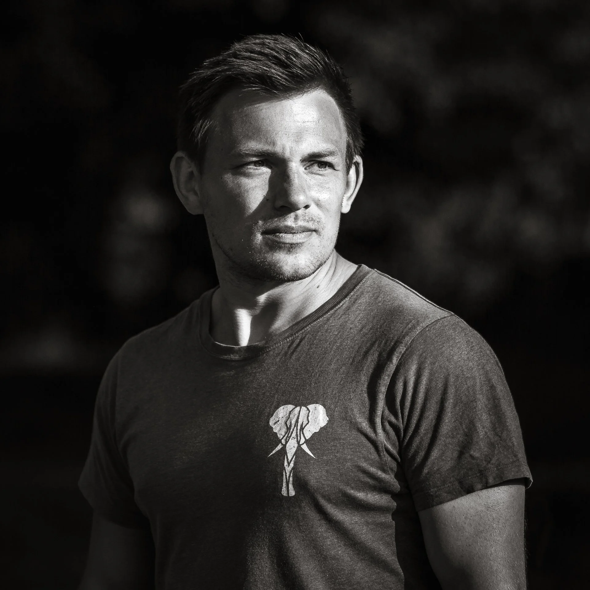 A young man with short hair looking to the right outdoors, wearing a t-shirt with an elephant graphic.