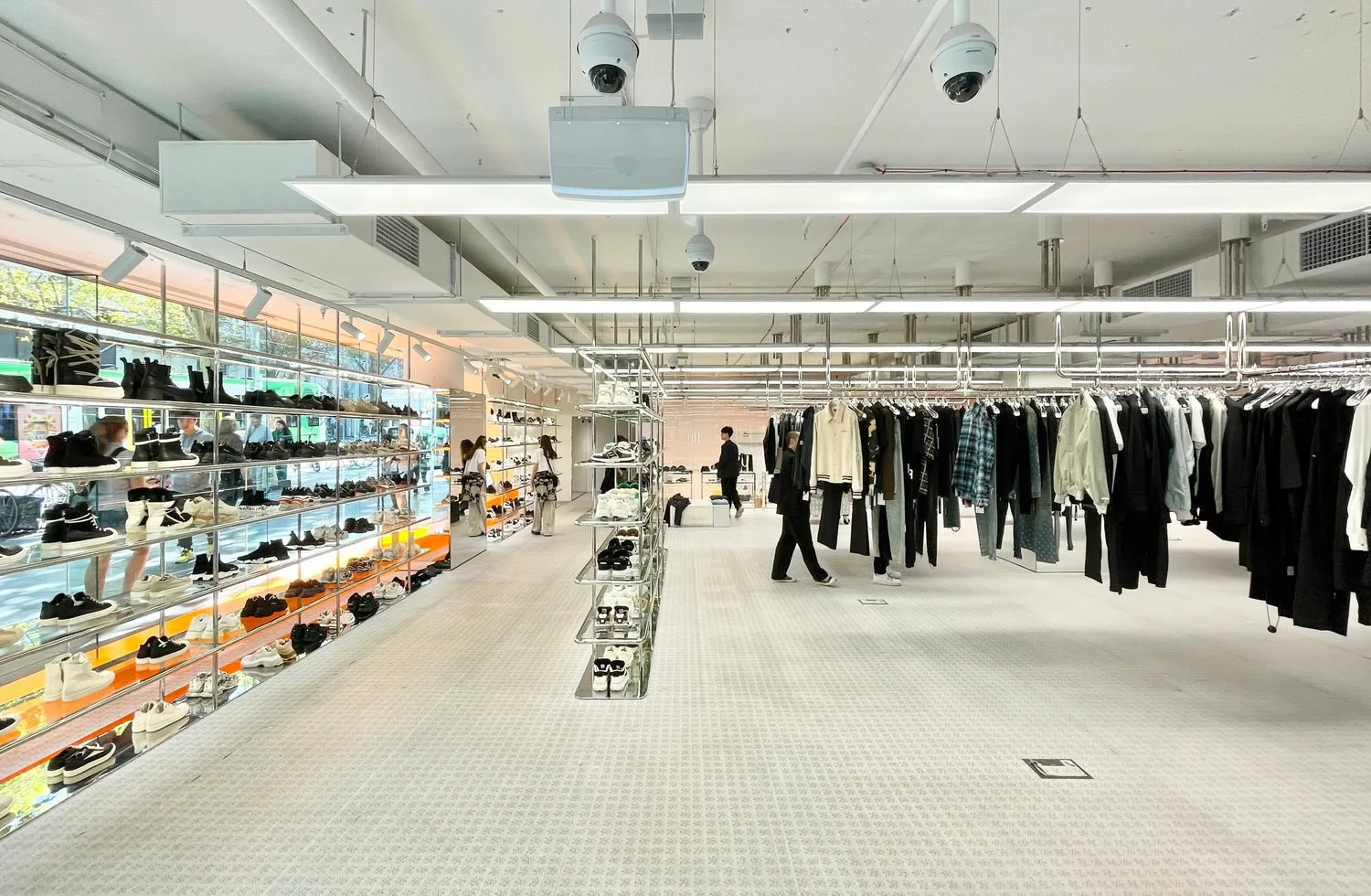 Interior of a clothing store featuring shoes on glass shelves on the left and hanging clothes on racks on the right. Several people are shopping and browsing inside the store.