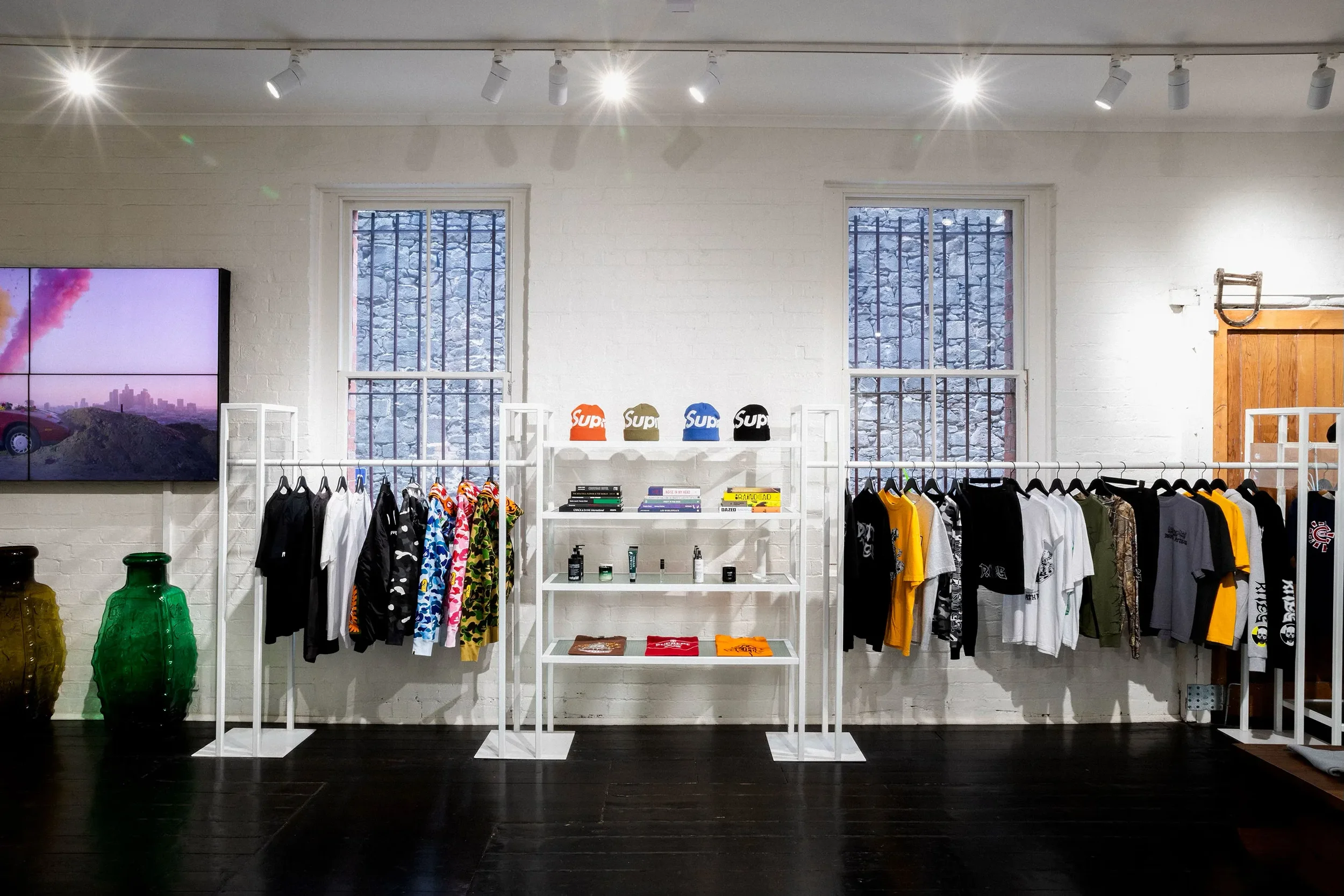 Clothing store interior with racks of T-shirts, hats on a shelf, and various decorative items against a white brick wall with two windows.