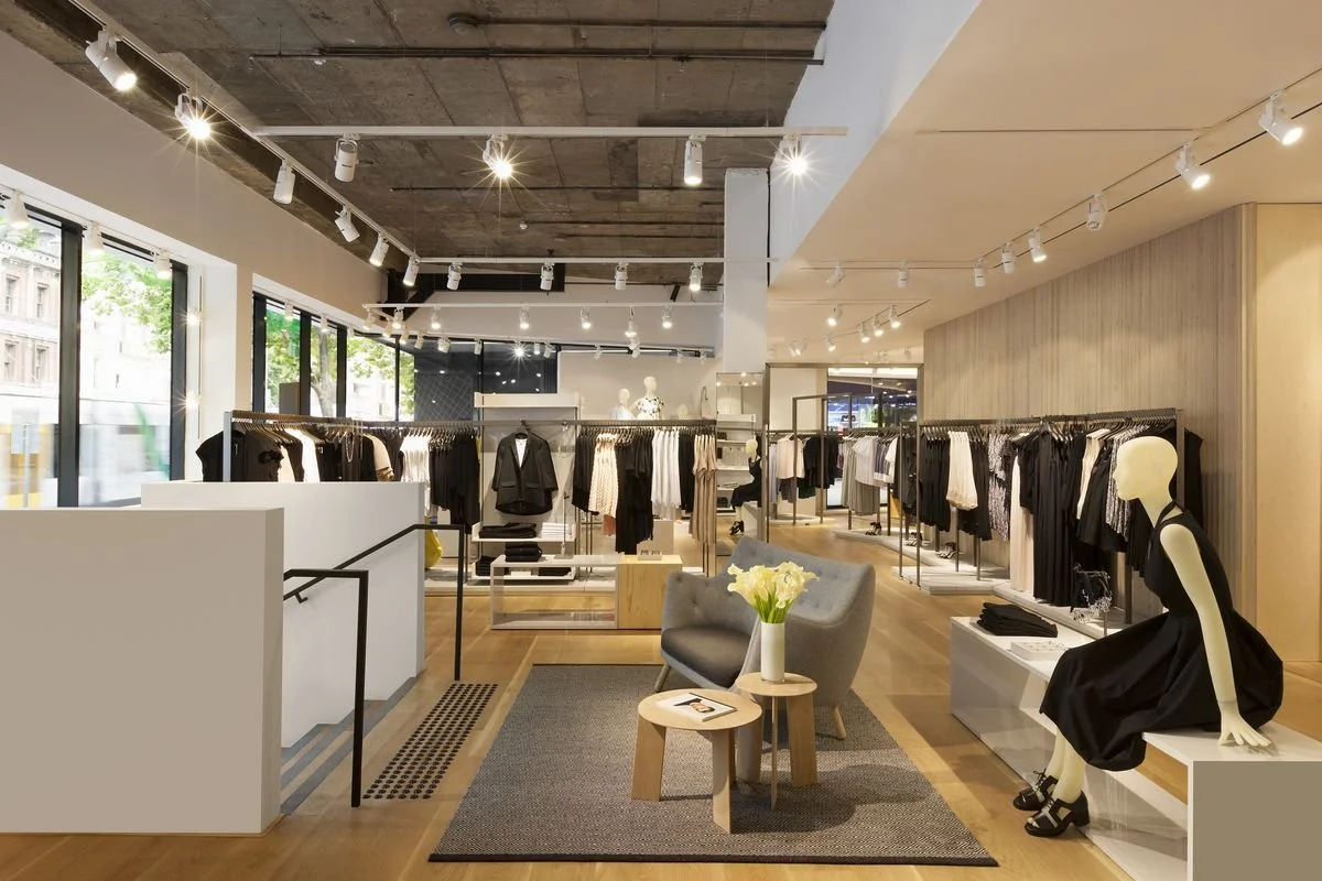 Interior of a clothing store with racks of clothing, a mannequin dressed in black, a gray armchair with a flower vase, and large windows letting in natural light.
