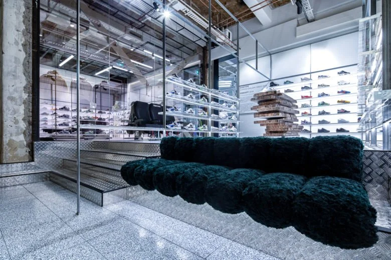 View of a luxury shoe store with glass walls, ceiling lights, and a black velvet sofa in the foreground.